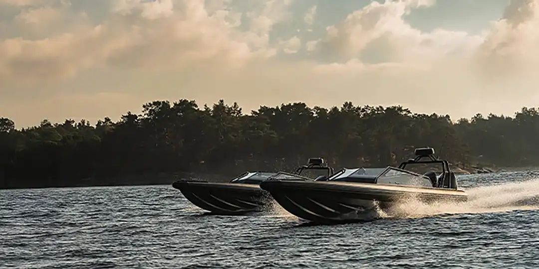For-utstallare-1080x540 Two fast motorboats run side by side on the water near a forested coast.