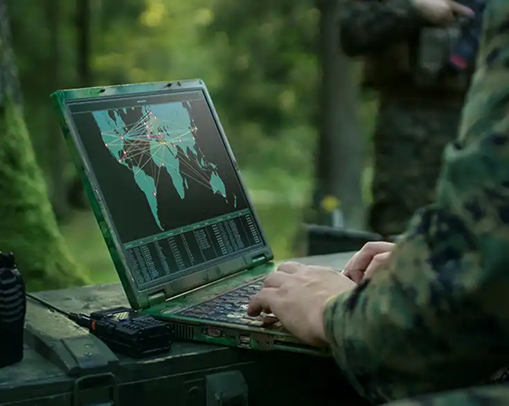 En person i militäruniform arbetar vid en laptop med en världskarta och dataströmmar på skärmen i en skogsmiljö.