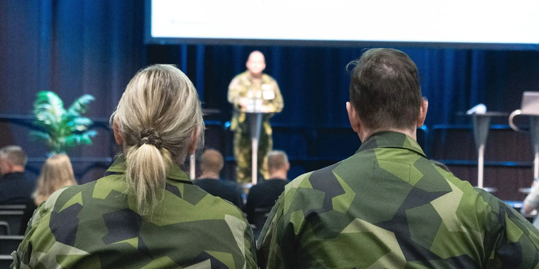 Deltagare i uniform sitter i publiken och lyssnar på en föreläsare på en scen Totalförsvarsmässan.  