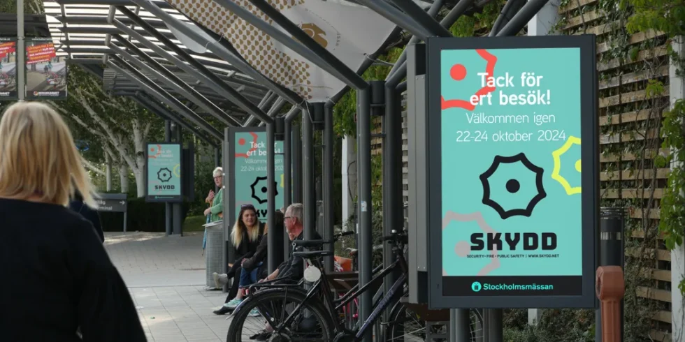 Outdoor digital screens along the walkway outside Stockholmsmässan displaying messages from the SKYDD exhibition, as visitors sit and pass through the area.