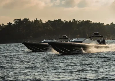 Two fast motorboats are traveling side by side over slightly wavy water. The water splashes up behind the boats as they travel at high speed. In the background, a forested coastline can be seen under a cloudy sky with warm, subdued light.