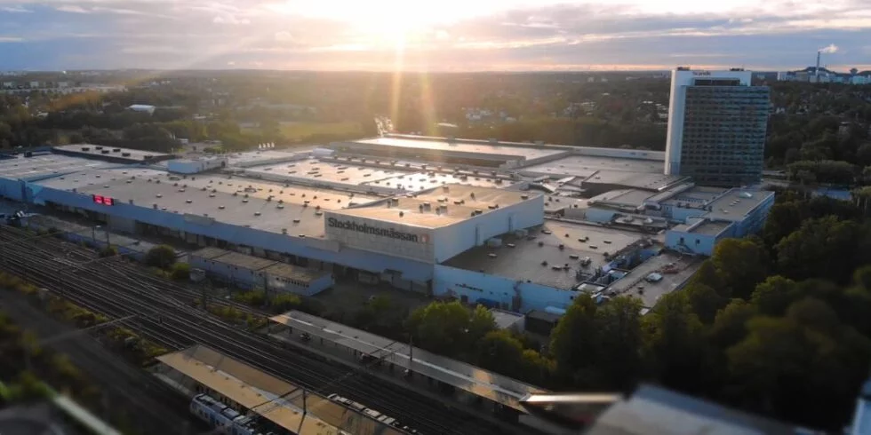 Stockholmsmassan_1080x540 Flygvy över Stockholmsmässan i Älvsjö i solnedgång, med mässhallarna, hotellet och tågspår i förgrunden.